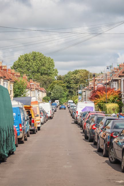 A residential street with rows of parked cars on both sides, including a blue van covered with a green tarp on the left and various other vehicles such as white vans and smaller cars on the right. The
