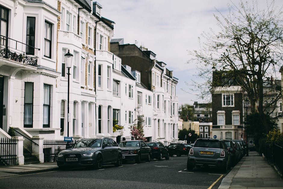 A residential street with rows of parked cars on both sides, including a blue van covered with a green tarp on the left and various other vehicles such as white vans and smaller cars on the right. The