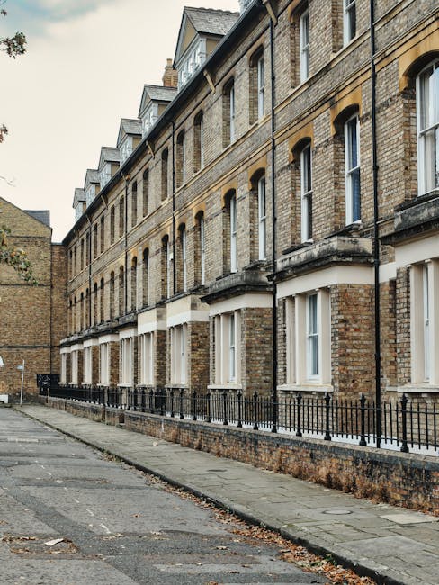 A row of traditional brick terraced houses with white-framed windows and small balconies, situated along a paved residential street in Brompton. The buildings are several stories high with slate roofs and chimneys, and a black metal fence runs parallel to the sidewalk in front of the houses. The pavement is made of concrete slabs, with some fallen leaves scattered along its surface. The scene is illuminated by natural daylight, highlighting the textures of the brickwork and the uniformity of the building facades. In the context of house removals, this image illustrates the exterior environment of terrace homes that require careful planning during furniture transport, home relocation, and loading processes, which Man and Van Brompton provides, particularly where narrow access and urban logistics are involved.