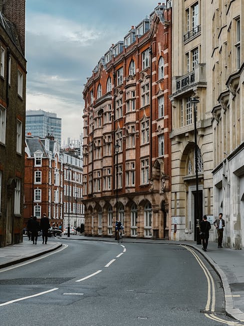 The image shows a narrow street on Old Brompton Road with a mix of historic and modern buildings, including a prominent multi-storey red-brick structure with ornate architectural details and white window frames. The street features a slightly curved road with double yellow lines along the kerb, indicating parking restrictions, and a white dashed centreline. On the pavement, several pedestrians are visible, some walking and others waiting near the sidewalk. There are a few bicycles parked against the building, and a person is seen loading or unloading items at the base of the brick building. The scene appears to be during daytime with natural, diffuse lighting under a cloudy sky. This setting captures the typical environment for house removals and furniture transport in urban London, where careful handling of belongings might involve moving through tight streets and negotiating urban building access, as managed by companies like Man and Van Brompton, specialists in home relocation services.