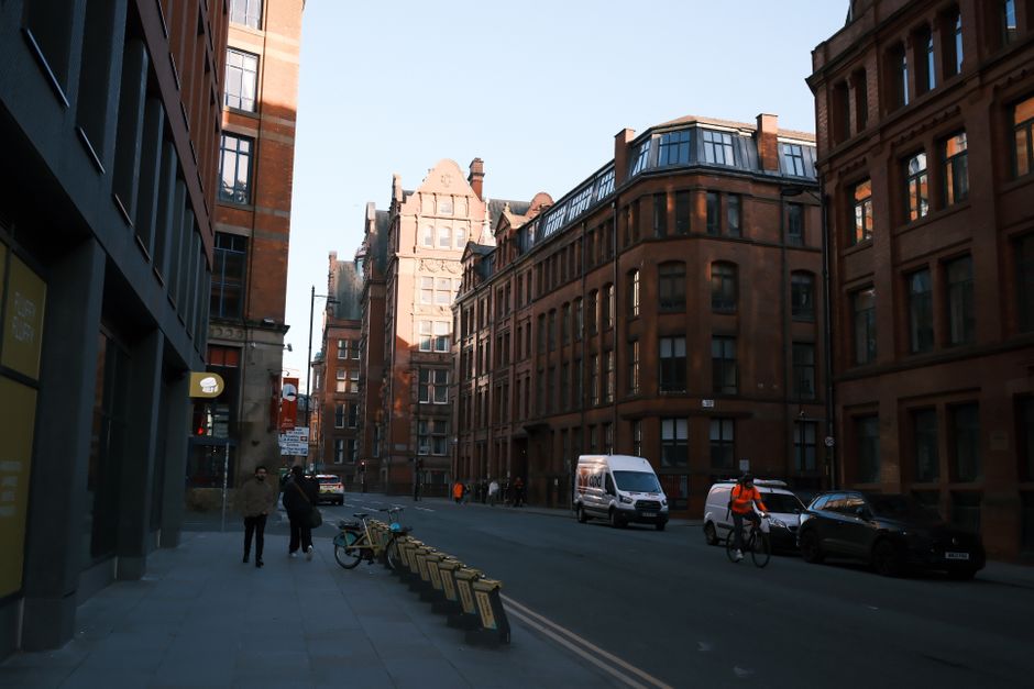 Photograph of a street scene on Old Brompton Road during daytime, featuring tall red-brick buildings with multiple windows and ornate architectural details. The street is lined with parked cars, including a white van in the process of loading or unloading, suggesting home relocation or furniture transport activity. A person in an orange safety vest is cycling along the road, while two pedestrians walk on the sidewalk. The scene includes various bins and bicycles secured to a bike rack on the pavement. Bright sunlight illuminates the buildings and street, with clear blue sky overhead. The presence of a moving van and the context of the street environment indicate ongoing packing or moving logistics, consistent with house removals services provided by Man and Van Brompton.