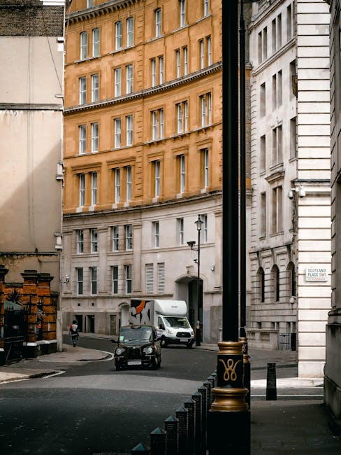 The image shows a narrow street on Old Brompton Road with a mix of historic and modern buildings, including a prominent multi-storey red-brick structure with ornate architectural details and white window frames. The street features a slightly curved road with double yellow lines along the kerb, indicating parking restrictions, and a white dashed centreline. On the pavement, several pedestrians are visible, some walking and others waiting near the sidewalk. There are a few bicycles parked against the building, and a person is seen loading or unloading items at the base of the brick building. The scene appears to be during daytime with natural, diffuse lighting under a cloudy sky. This setting captures the typical environment for house removals and furniture transport in urban London, where careful handling of belongings might involve moving through tight streets and negotiating urban building access, as managed by companies like Man and Van Brompton, specialists in home relocation services.