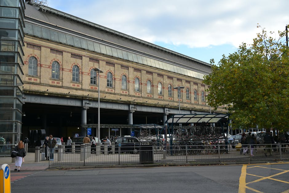 A modern passenger train with a sleek silver and white exterior, accented by orange and black details, is stationary at West Brompton station platform. The train's front end features a large, tinted w